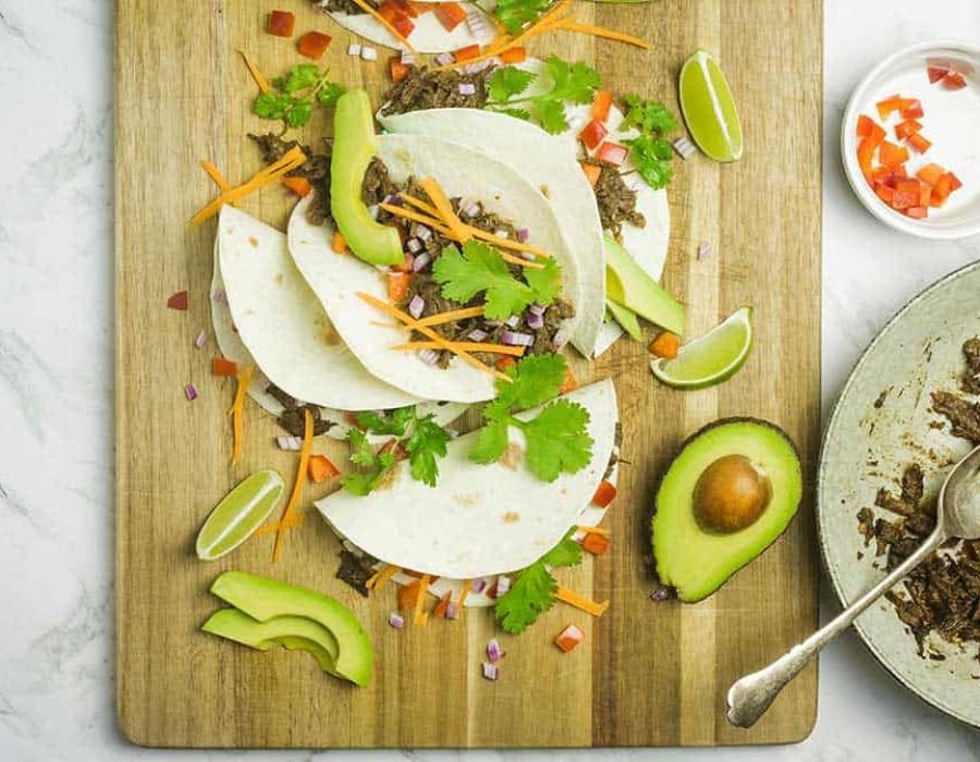 Close up view of soft tacos with shredded beef, julienned carrot, diced onion & tomato on a wooden board on a marble countertop.