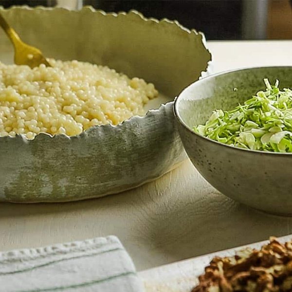 Rustic earthenware bowls with pearl couscous and shredded Brussels sprouts, alongside a dish with toasted pecans and dried cranberries.