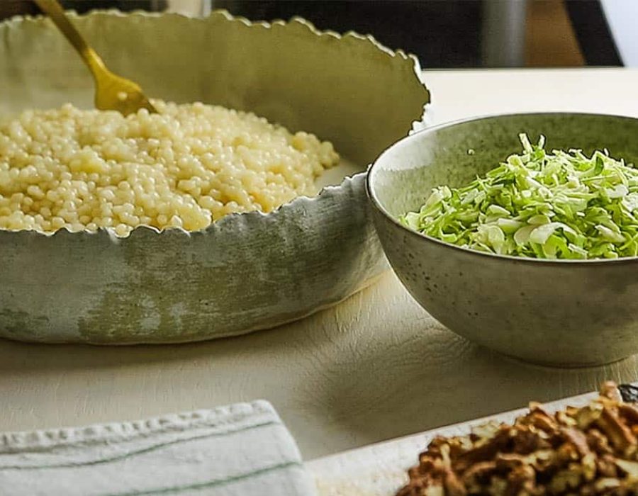 Rustic earthenware bowls with pearl couscous and shredded Brussels sprouts, alongside a dish with toasted pecans and dried cranberries.