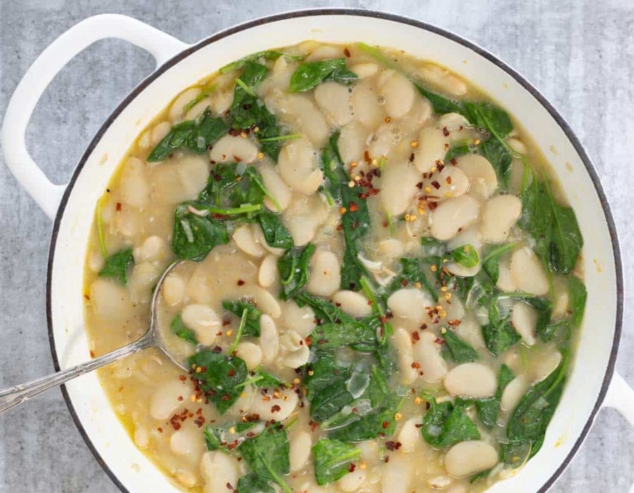 A large white cast iron pot is viewed from above. In the pot is creamy butter beans with lemon, garlic and spinach. Sprinkled on top is a scattering of red pepper flakes.
