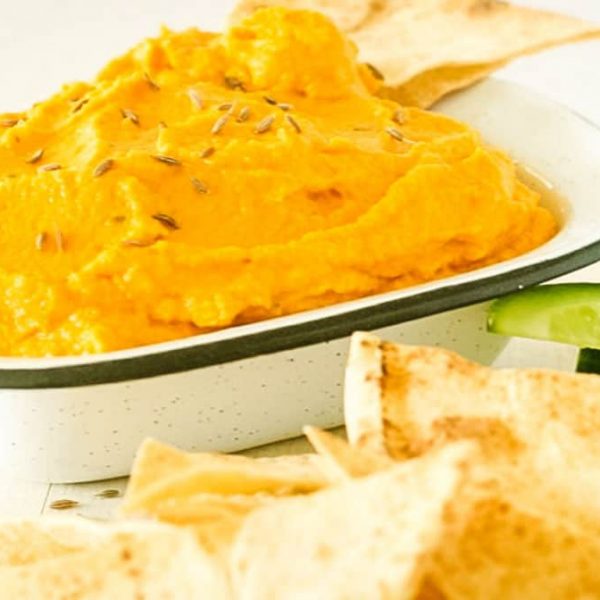 A speckled dish with carrot hummus alongside tortilla chips and cucumber strips.
