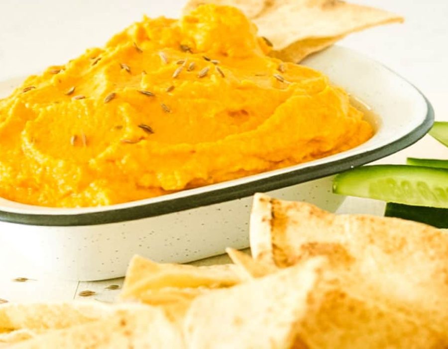 A speckled dish with carrot hummus alongside tortilla chips and cucumber strips.