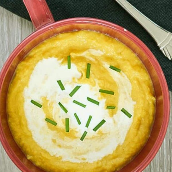 A red soup mug with thick, golden cauliflower and pumpkin soup garnished with chives