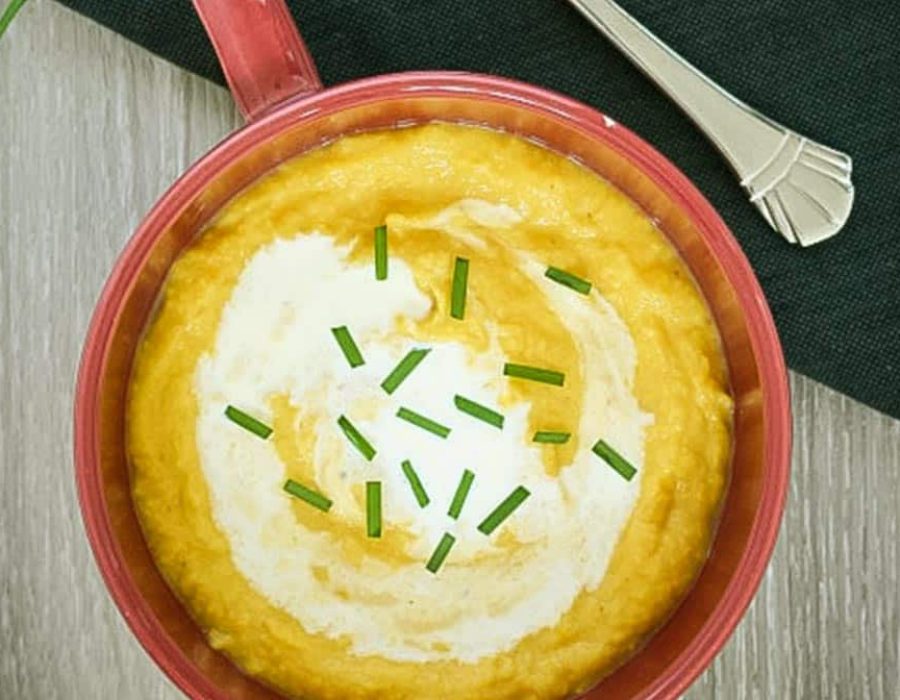 A red soup mug with thick, golden cauliflower and pumpkin soup garnished with chives