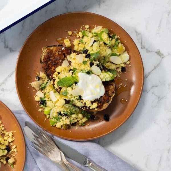 Landscape view of a terracotta plate holding a roasted eggplant half topped with yellow couscous, sliced green chilies, yogurt, and a drizzle of honey on a marble surface.