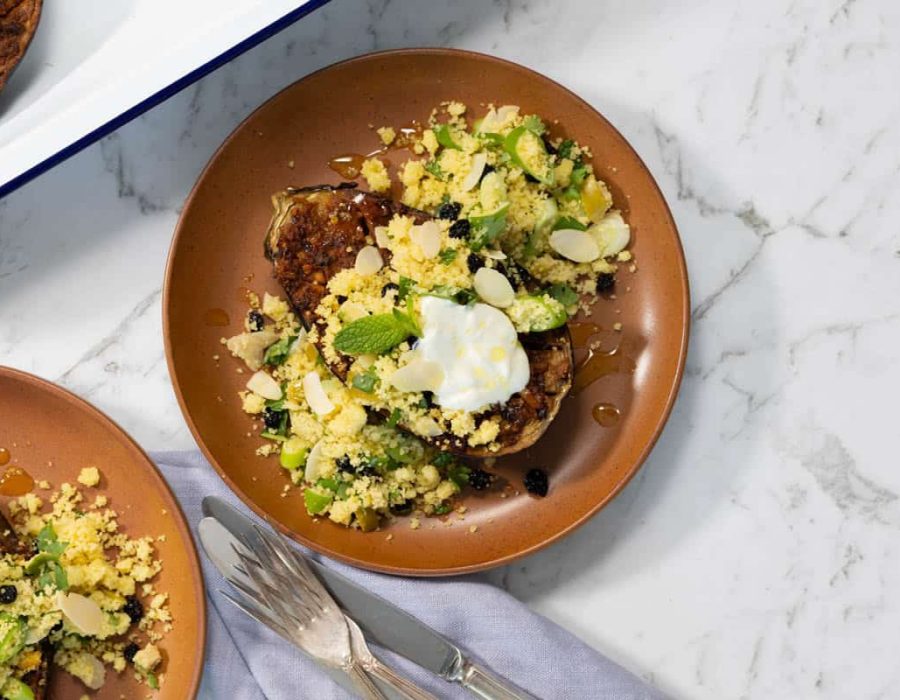 Landscape view of a terracotta plate holding a roasted eggplant half topped with yellow couscous, sliced green chilies, yogurt, and a drizzle of honey on a marble surface.