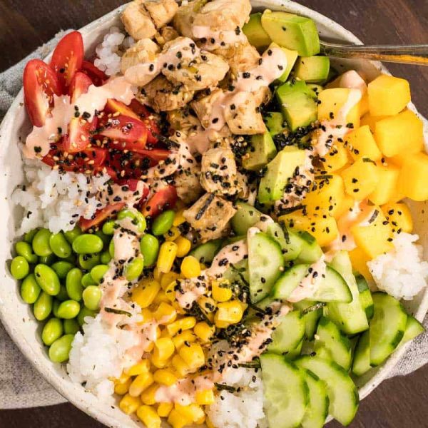 A full to overflowing bowl of sushi rice, steamed chicken, tomatoes, avocado, corn, mango, cucumber and edamame beans. The bowl is topped with spicy mayo and furikake seasoning to create a chicken poke bowl.