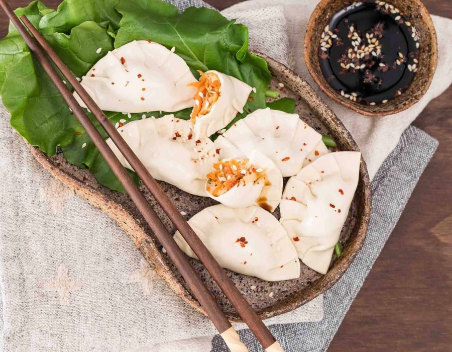 A kidney shaped brown ceramic plate with 6 steamed chicken dumplings and steamed broccoli greens arranged on it. One of the dumplings has been broken open to reveal chicken and carrot filling, and a set of dark wooden chopsticks lays across the plate.