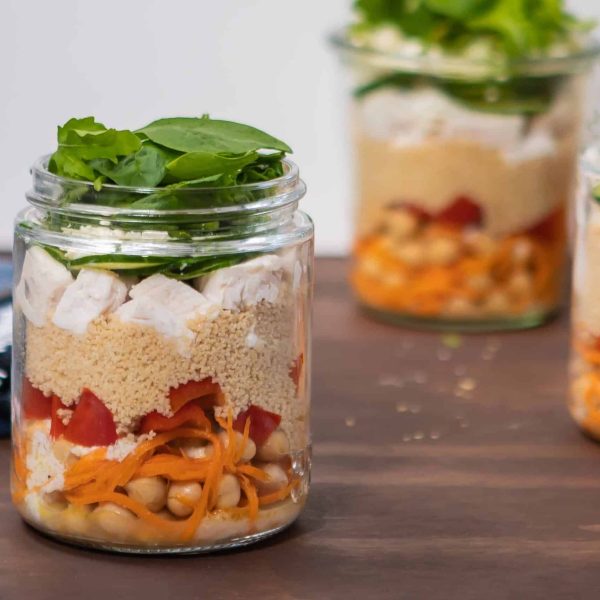 a landscape image of a jar of chickpea chicken salad, with baby spinach piled into the top of the jar and spilling over the sides