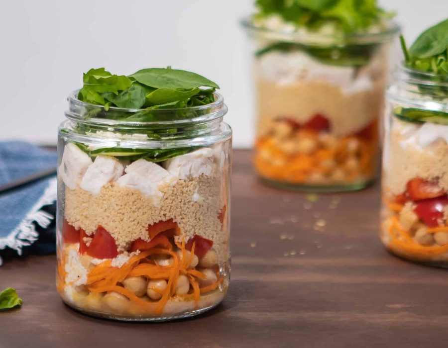 a landscape image of a jar of chickpea chicken salad, with baby spinach piled into the top of the jar and spilling over the sides