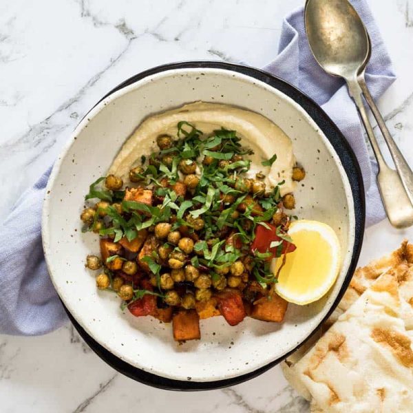 shallow ceramic bowl lined with hummus and topped with roasted sweet potatoes, peppers, crispy chickpeas, fresh chopped parsley, and a lemon wedge alongside pita bread.