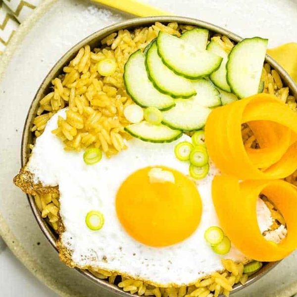 A bowl with seasoned rice topped with a fried egg, cucumber moons, pickled carrot and scallions