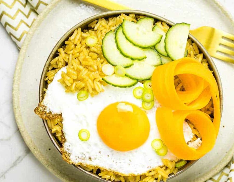 A bowl with seasoned rice topped with a fried egg, cucumber moons, pickled carrot and scallions