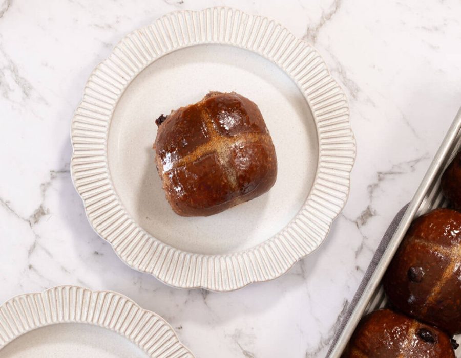 A single chocolate milk bread hot cross bun, on a white scalloped plate. The remaining tray of buns can be seen at the edge of frame.