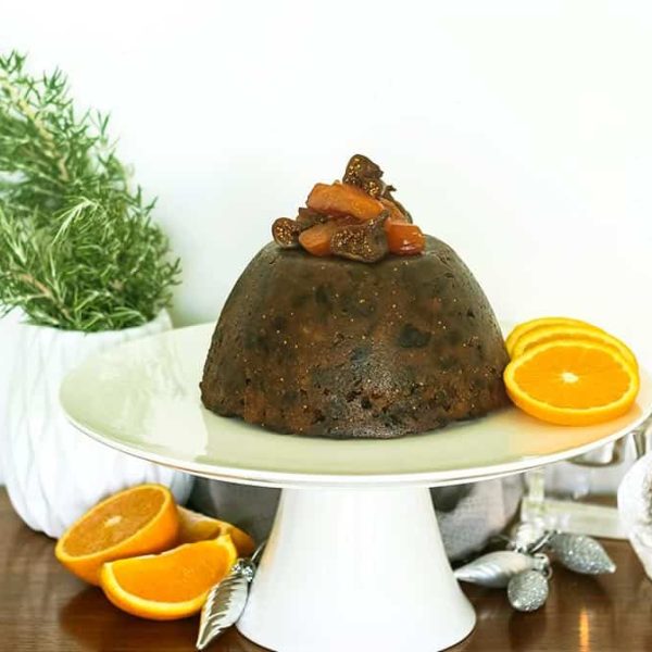 a Christmas pudding garnished with fruit on a white cake stand surrounded by citrus and decorations
