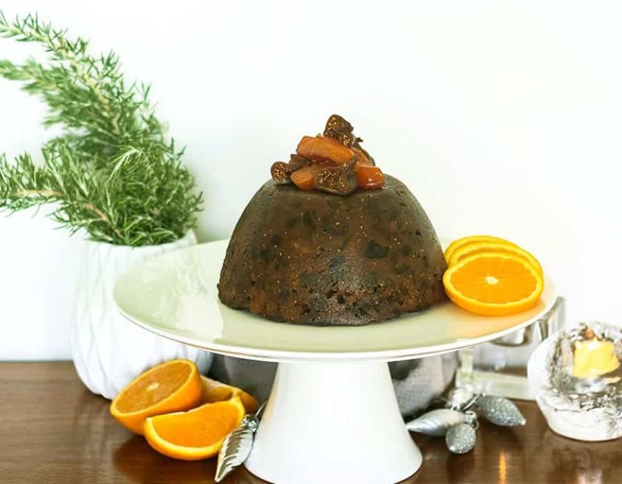 a Christmas pudding garnished with fruit on a white cake stand surrounded by citrus and decorations