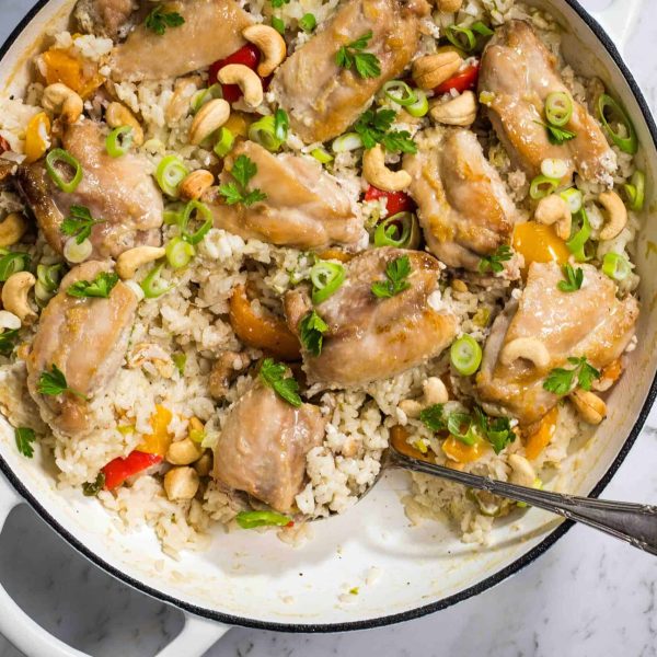 a top down view of a white casserole dish with cooked rice, chicken thighs and bell peppers, topped with cashew nuts and scallions