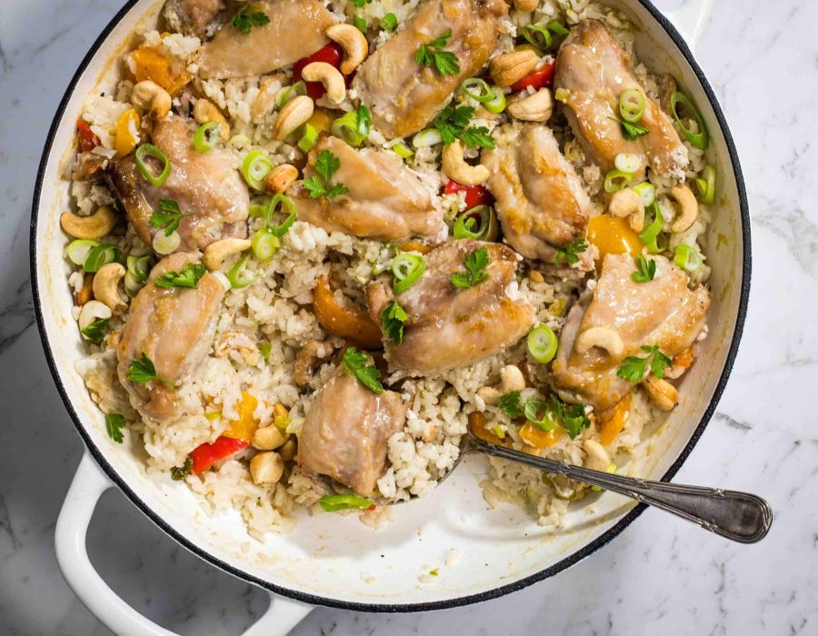 a top down view of a white casserole dish with cooked rice, chicken thighs and bell peppers, topped with cashew nuts and scallions