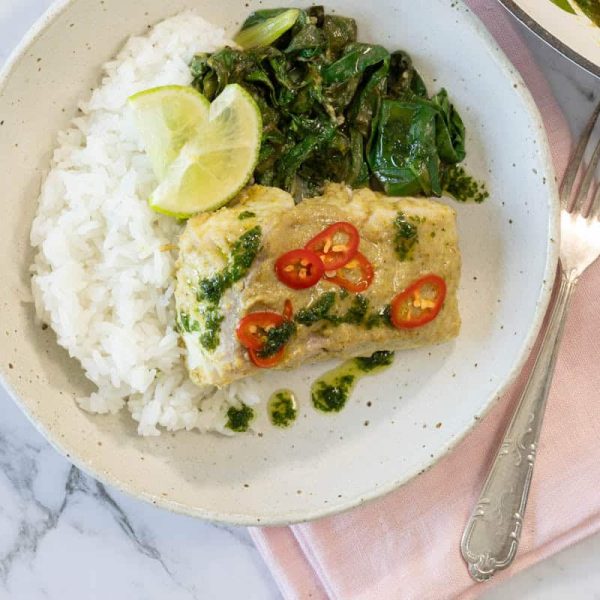 Overhead landscape shot of a ceramic bowl filled with coconut curry steamed fish, rice, Swiss chard, and lime with a fork on a pink napkin for coconut steamed fish.