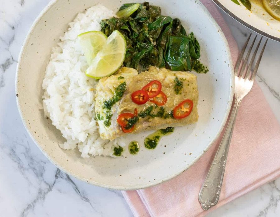 Overhead landscape shot of a ceramic bowl filled with coconut curry steamed fish, rice, Swiss chard, and lime with a fork on a pink napkin for coconut steamed fish.