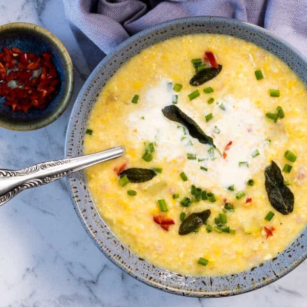 A close-up, overhead view of a blue ceramic bowl filled with corn and bacon chowder as the sour cream starts to swirl into the broth, topped with fried sage leaves and green onions next to a silver spoon and a small dish of diced red peppers for corn and bacon chowder.