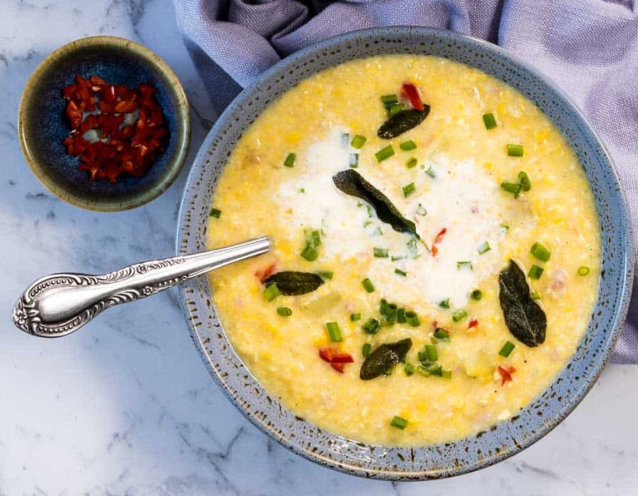 A close-up, overhead view of a blue ceramic bowl filled with corn and bacon chowder as the sour cream starts to swirl into the broth, topped with fried sage leaves and green onions next to a silver spoon and a small dish of diced red peppers for corn and bacon chowder.