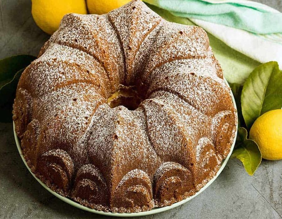 A whole cream cheese pound cake, turned out of a decorative bundt pan and dusted with icing sugar. A lemon and a green tea towel sit to the side.
