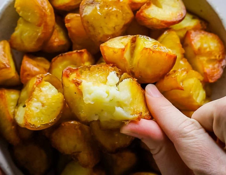 Two fingers hold a crispy roasted potato up to the camera from below. The potato has been broken open to show the crispy exterior and the fluffy, soft interior. Underneath the potato a bowl of roasted potatoes can be seen slightly out of focus.
