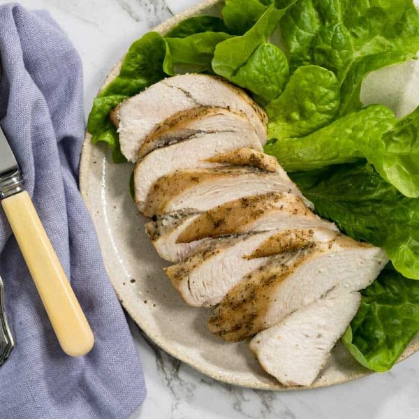Slices of juicy roasted chicken breast are arranged on a light ceramic plate alongside fresh green butter lettuce leaves, complete with a silver fork and knife on a blue cloth napkin for dry brined chicken.