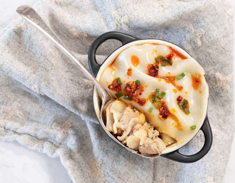 Overhead view of a cooked dumpling lasagne in a small black-handled cocotte topped with chili crisp and sliced green onions on a blue patterned cloth.