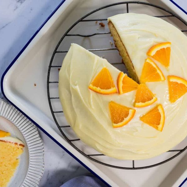 Sliced sponge cake topped with smooth white frosting and fresh orange wedges sits on a wire rack next to a single serving on a fluted plate for easy orange cake.
