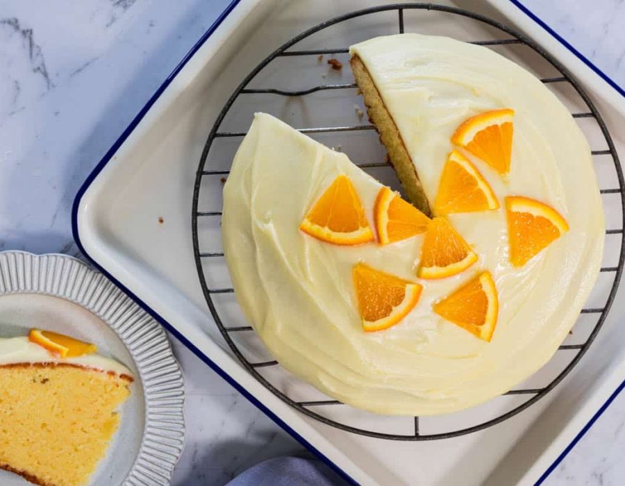 Sliced sponge cake topped with smooth white frosting and fresh orange wedges sits on a wire rack next to a single serving on a fluted plate for easy orange cake.