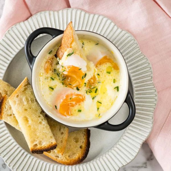steam-baked eggs topped with chives in a small black ramekin, served with toasted sourdough soldiers on a ribbed plate alongside a pink napkin.