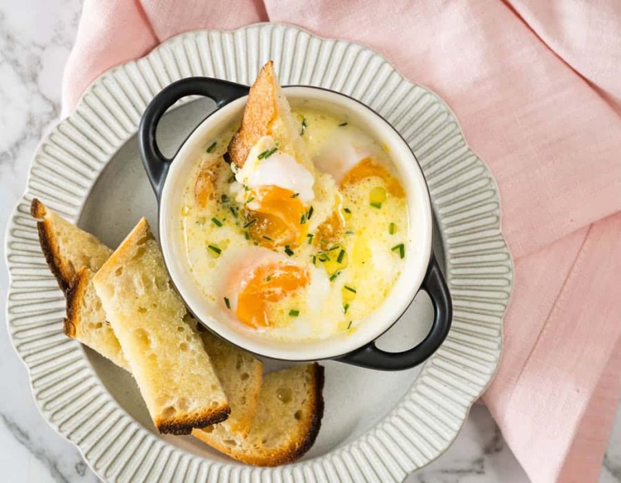 steam-baked eggs topped with chives in a small black ramekin, served with toasted sourdough soldiers on a ribbed plate alongside a pink napkin.