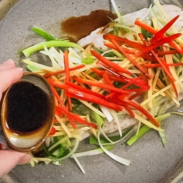 a hand holds a small bowl of soy sauce, drizzling it over the top of a plate containing steamed white fish, chilli, scallions and ginger
