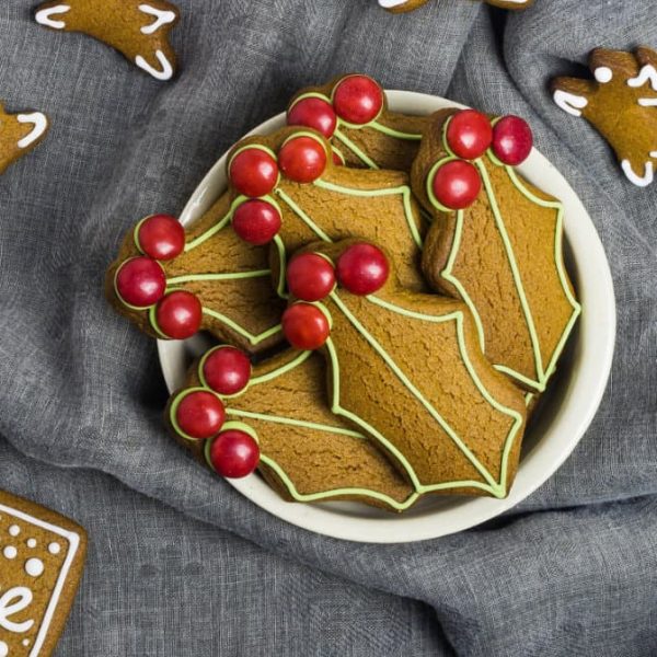 a white bowl filled with holly shaped gingerbread cookies with reindeer biscuits on a grey cloth