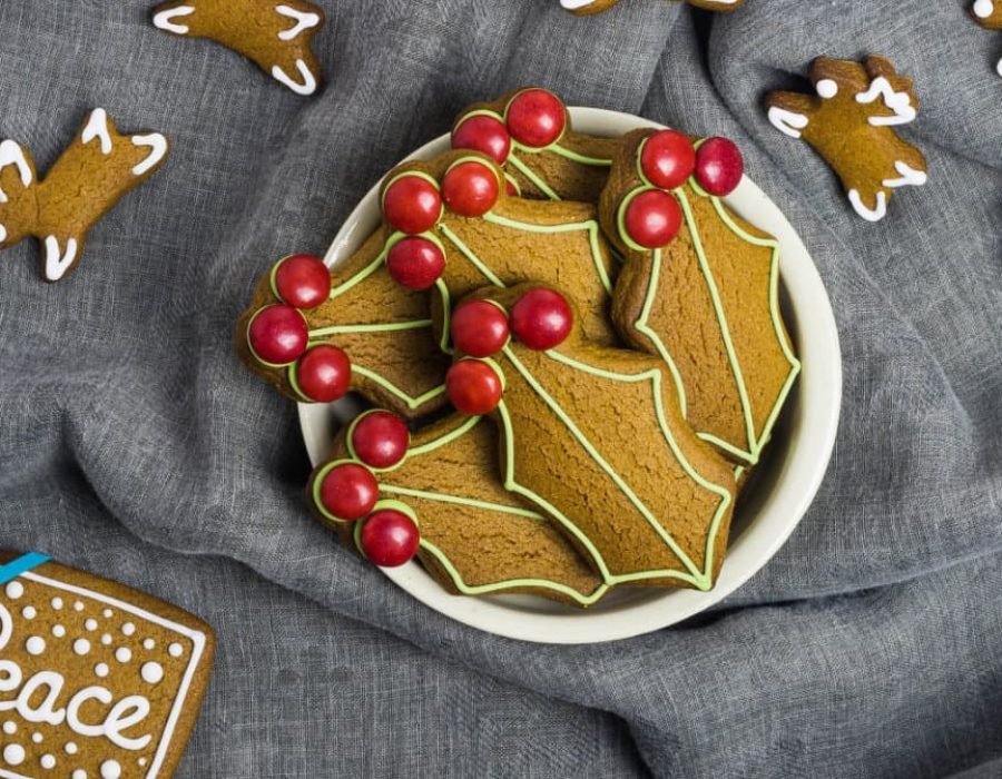 a white bowl filled with holly shaped gingerbread cookies with reindeer biscuits on a grey cloth