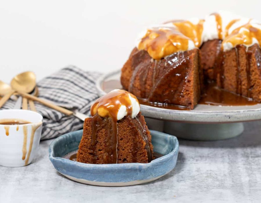 A sticky gingerbread steamed pudding on a large footed plate. The pudding has a piece cut out which can be seen on a plate in front; butterscotch sauce and whipped cream garnish the pudding.