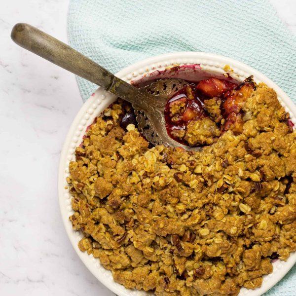 a white baking dish with a decorative serving spoon, with a berry coloured fruit crumble on a mint green tea towel