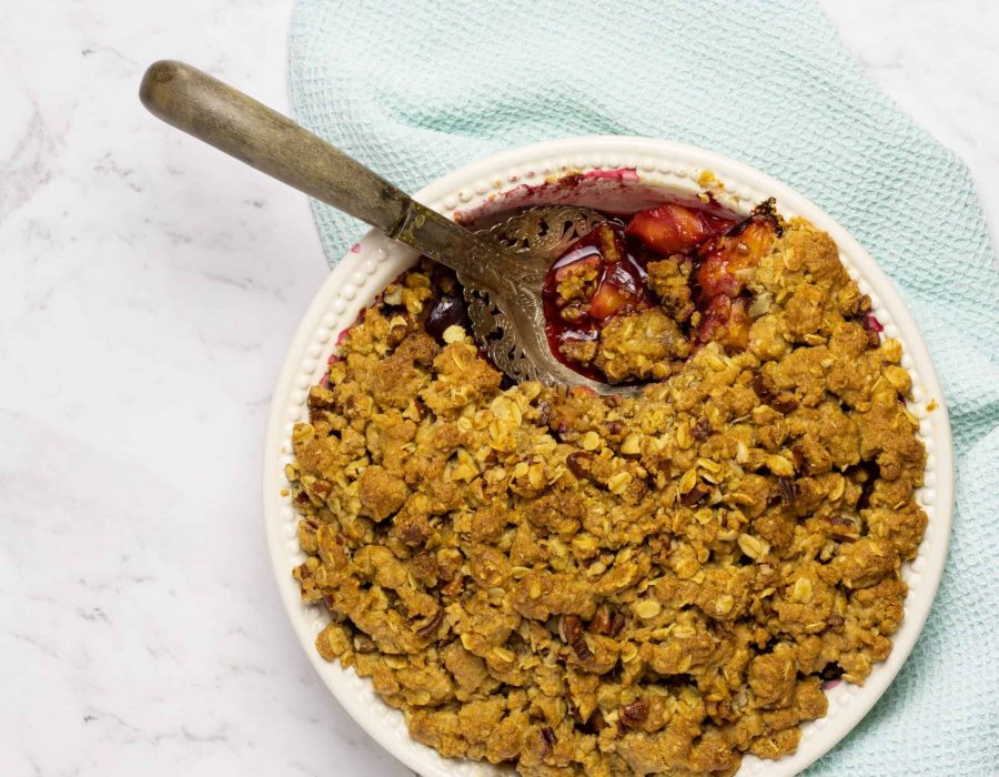 a white baking dish with a decorative serving spoon, with a berry coloured fruit crumble on a mint green tea towel