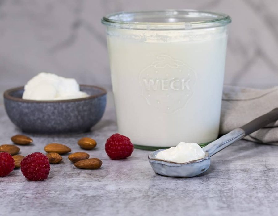 A small grey bowl with plain yoghurt in it; next to the bowl is a larger glass jar of yoghurt, and a ceramic spoon. There are almonds and raspberries scattered on the counter nearby.