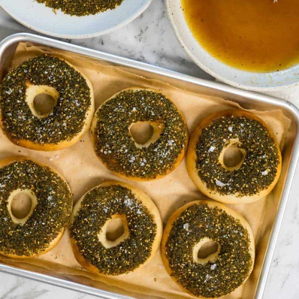 Six golden bagels topped with a thick layer of green zaatar herbs and sea salt flakes are arranged on a parchment-lined baking sheet for zaatar bagels.