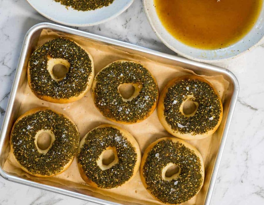 Six golden bagels topped with a thick layer of green zaatar herbs and sea salt flakes are arranged on a parchment-lined baking sheet for zaatar bagels.