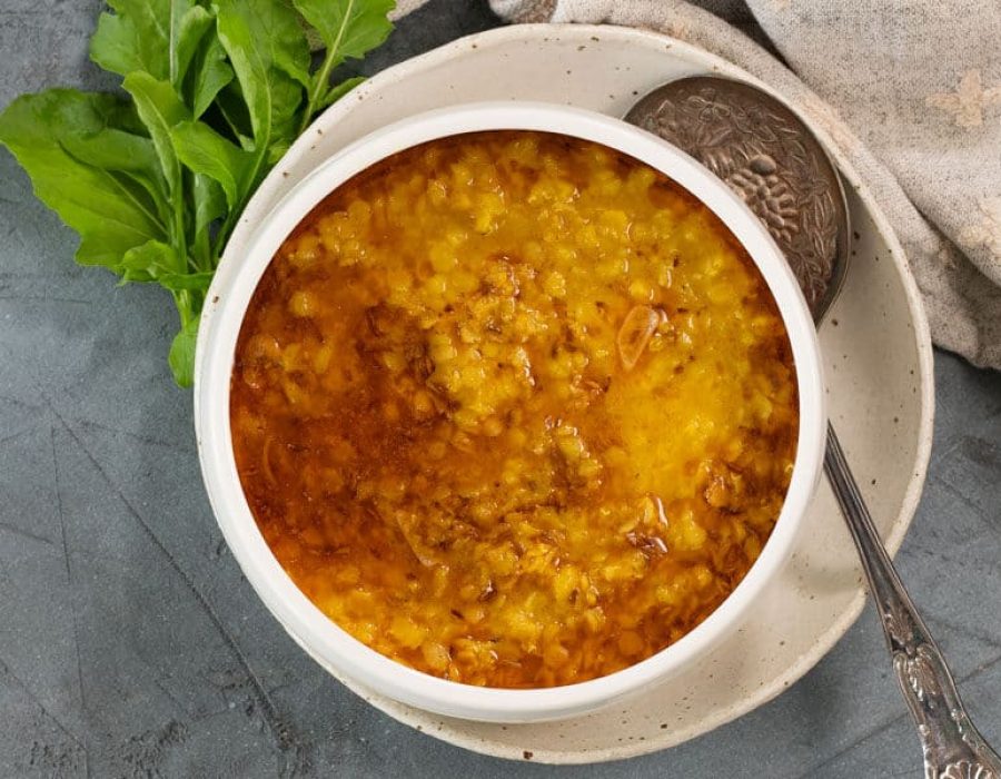 a white ceramic bowl of yellow and orange red lentil dahl, viewed from above, with a silver serving spoon, cloth napkin and leafy greens pictured alongside the bowl.