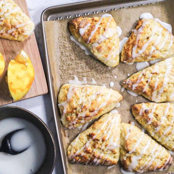 A gold baking sheet with parchment paper and 7 freshly baked triangular lemon scones with lemon glaze on it. Another scone sits to the side on a wooden board.