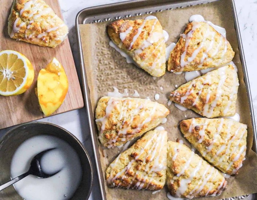 A gold baking sheet with parchment paper and 7 freshly baked triangular lemon scones with lemon glaze on it. Another scone sits to the side on a wooden board.