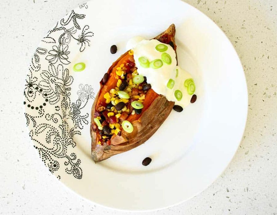 A loaded sweet potato stuffed with beans and corn, topped with sour cream garnished with sliced green onions on a patterned plate.