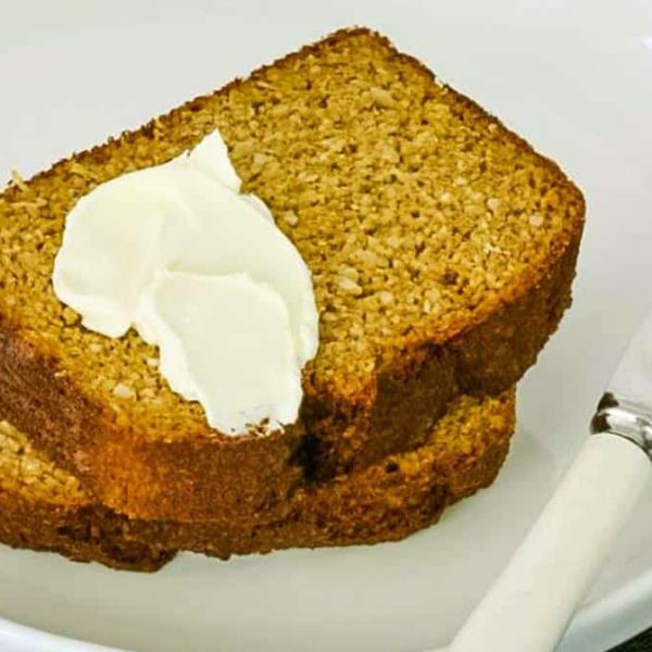 zoomed in image of two thick slices of pumpkin bread with a blob of cream cheese on top and a knife beside them on a white plate