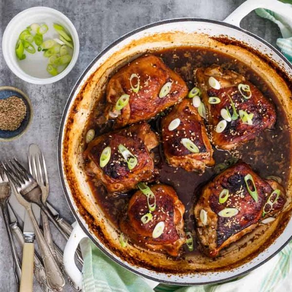 A round white cast iron baking dish is viewed from above. In the dish are 6 roasted chicken cutlets with miso maple marinade. There are sesame seeds and scallions sprinkled on top, and next to the dish is a pile of cutlery and extra scallions and seeds.