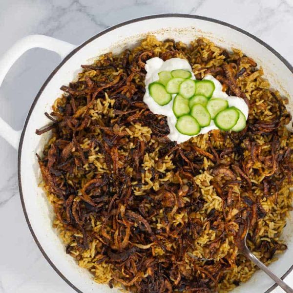 Mujadara-Lentil-Rice-process-10 Lentil rice served in a white pot, topped with crispy onions, yogurt, and cucumber slices.
